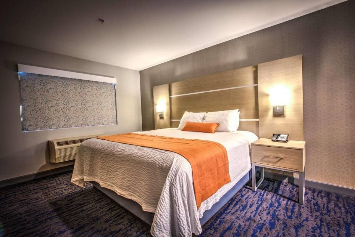 A hotel room with a large bed, a nightstand with a phone, a wall-mounted TV, and a window with blinds.