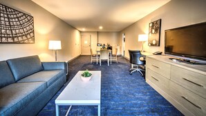 Une chambre d’hôtel avec un canapé, une table basse agrémentée d’une plante, un téléviseur, un bureau avec une chaise, et un coin cuisine à l’arrière-plan.
