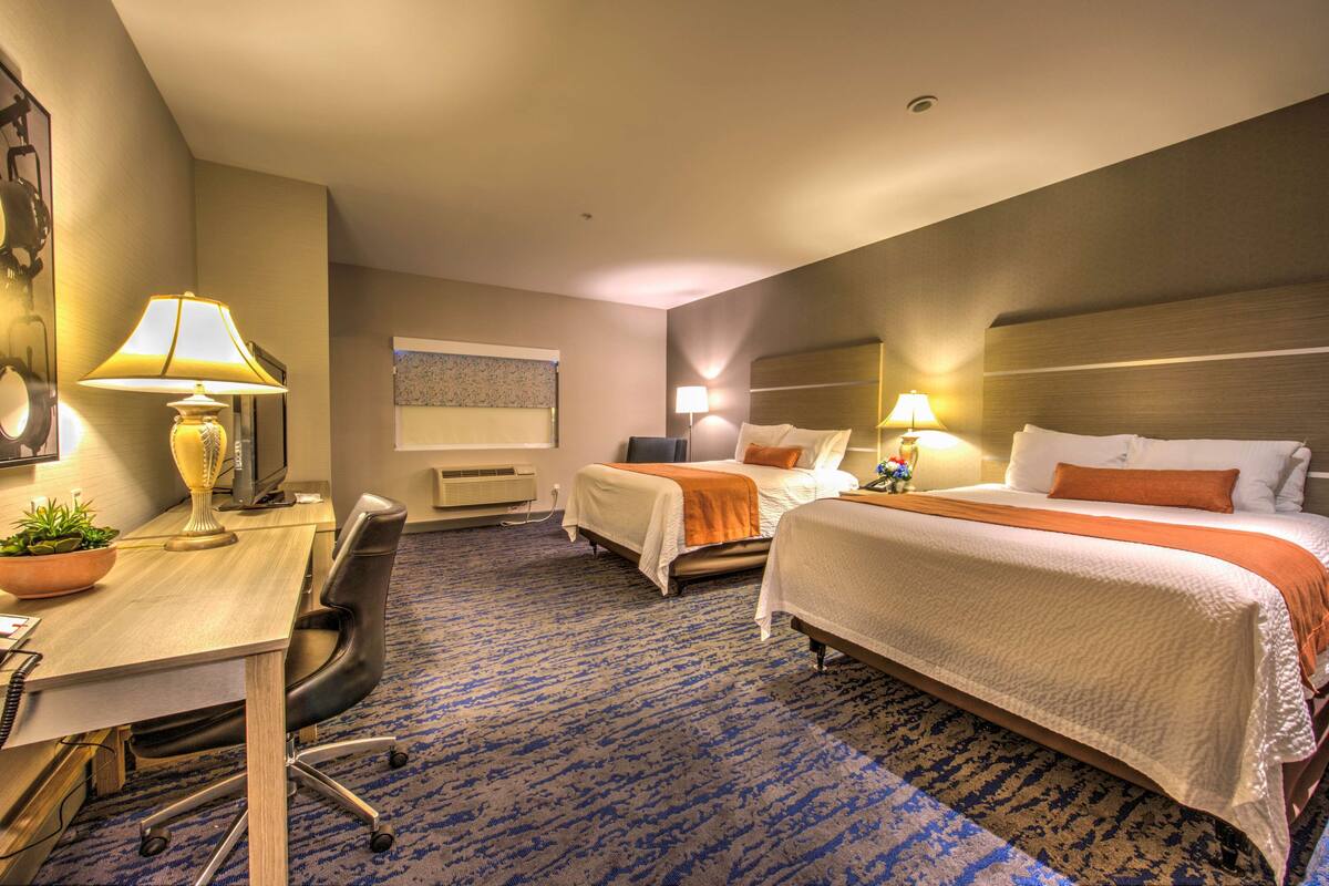 A hotel room with two beds, a desk, a chair, and a lamp.