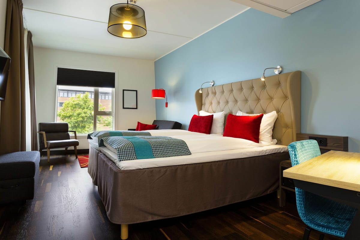 A hotel room with a large bed, a desk, a chair, a TV, and a window with a view of buildings and trees.