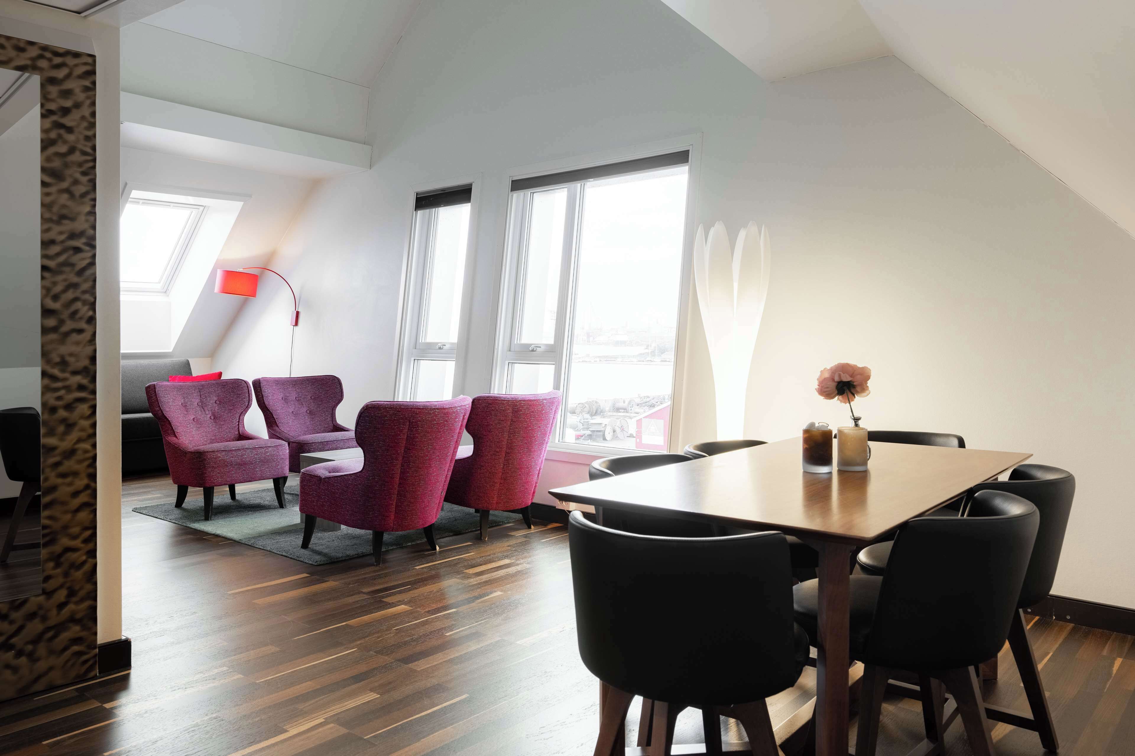 A modern dining area with a wooden table and black chairs, a red floor lamp, and a view through large windows.