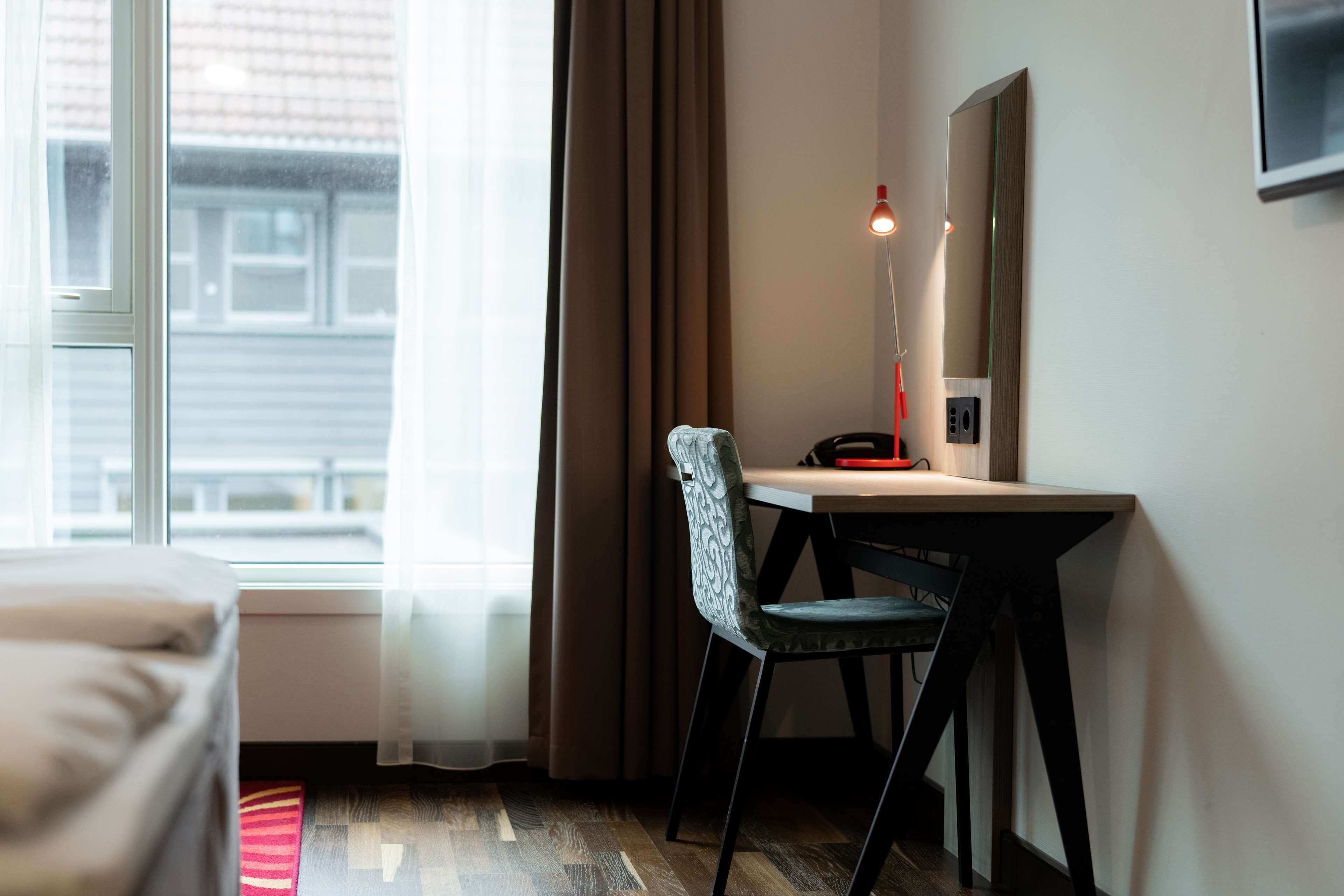 Quarto de hotel com cama, escrivaninha com cadeira, luminária e janela com cortinas.