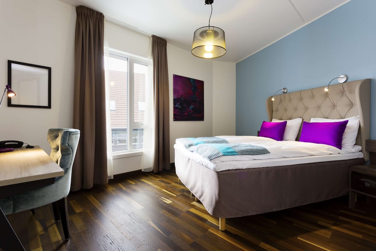 A bedroom with a bed, a desk, a chair, a window, and a painting on the wall.