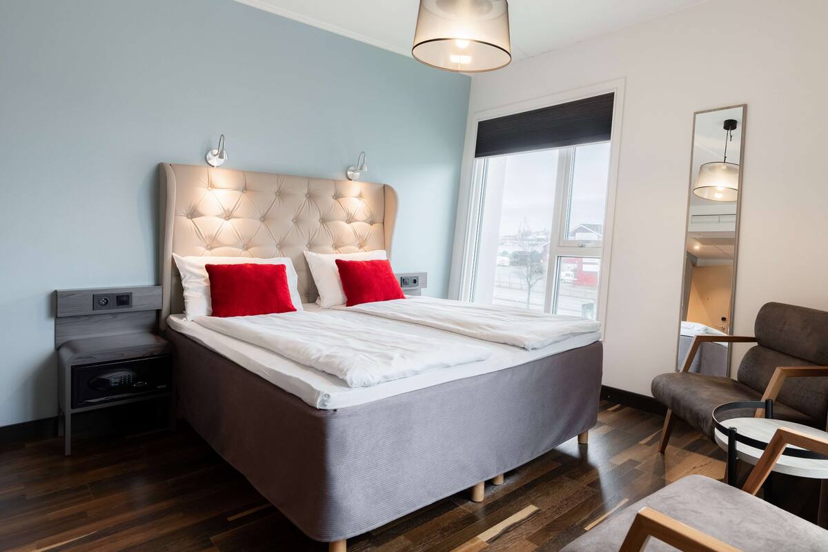 A modern bedroom with a tufted headboard, a bed with white and red bedding, a bedside table, a chair, and a large mirror.
