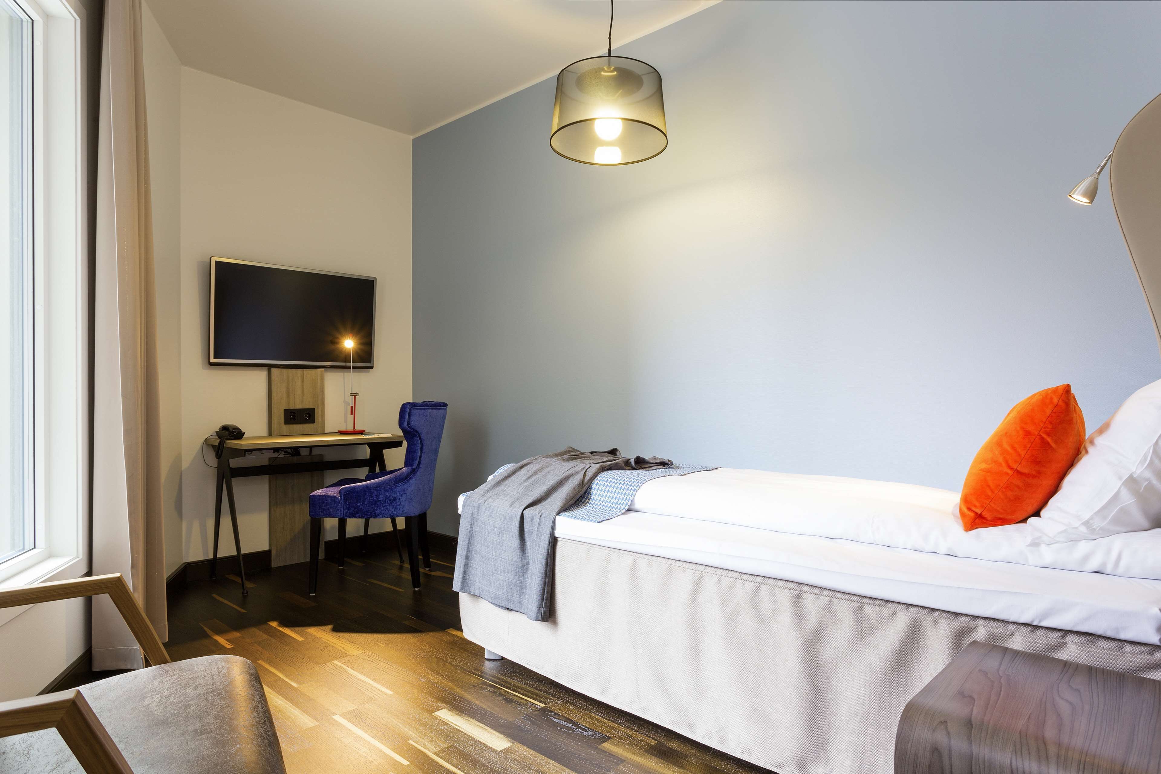 A modern bedroom with a bed, a desk with a chair, a television, and a hanging light fixture.