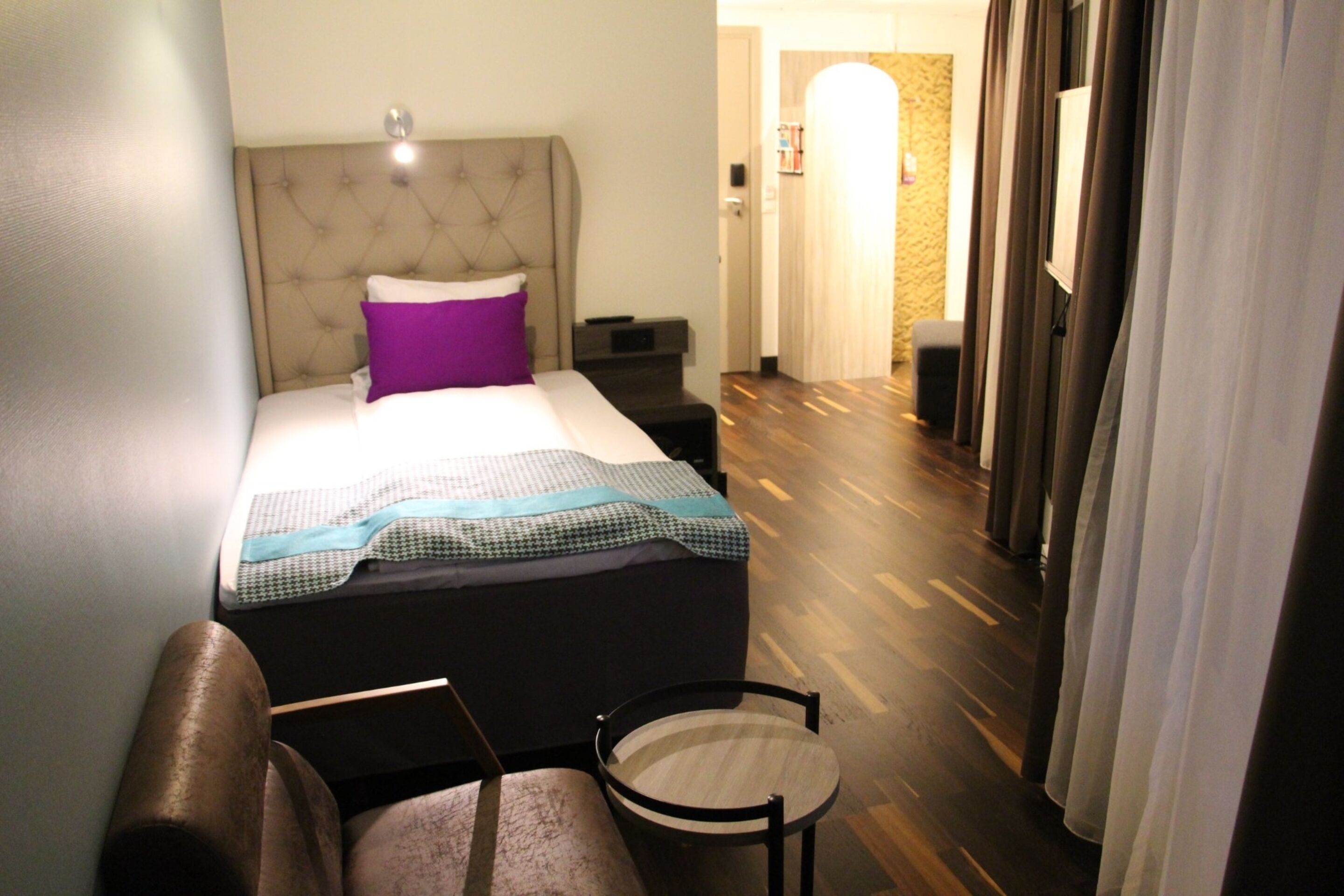 Una habitación de hotel con una cama, una silla, una mesita de noche y suelo de madera.