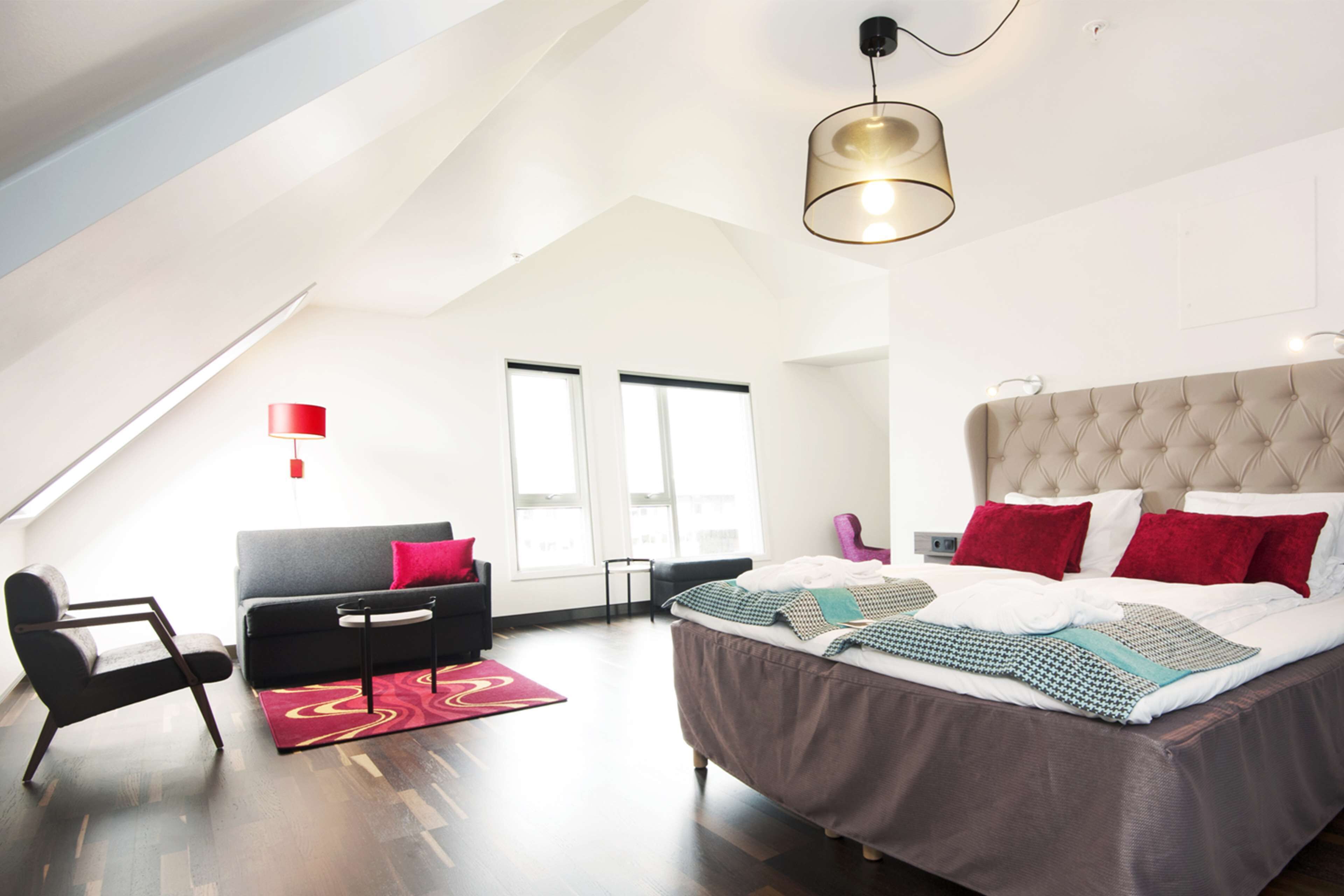 Un dormitorio moderno con una cama grande, un sofá, una silla y un escritorio. Hay dos ventanas y una lámpara en el techo.