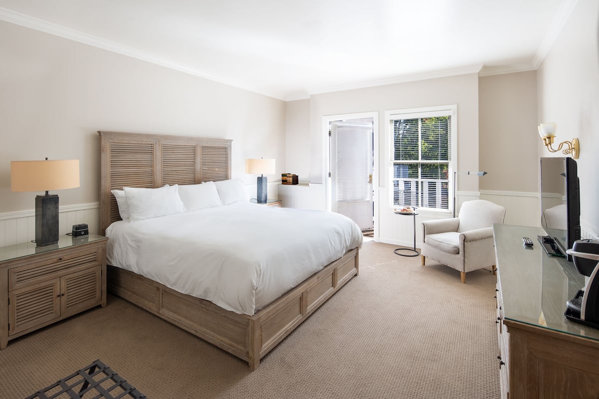 A bedroom with a large bed, bedside tables, a chair, a desk with a TV, and a window with blinds.