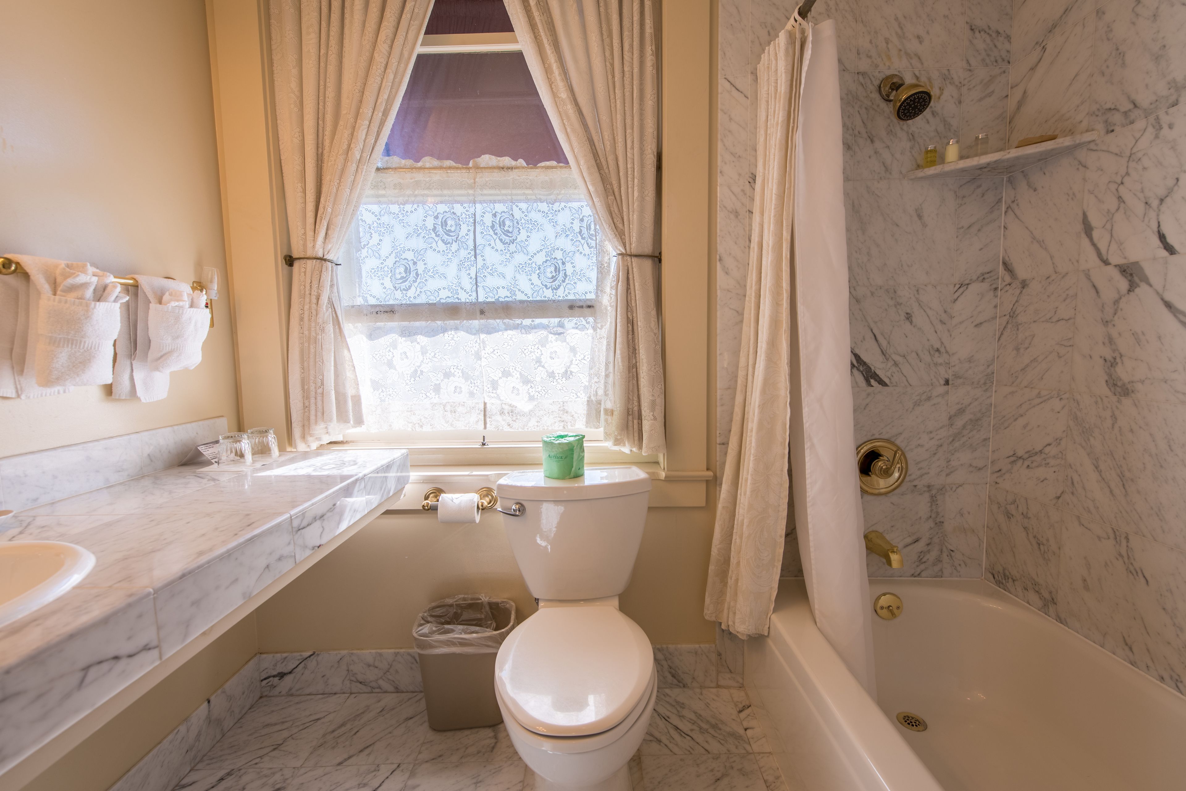 Een badkamer met toilet, wastafel en bad. Er is een raam met gordijnen, een prullenbak en een douchegordijn.