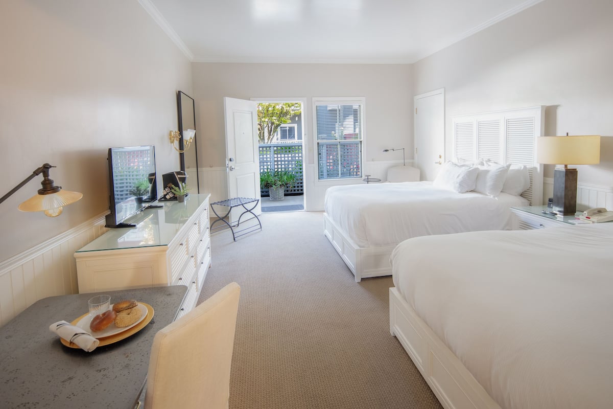 A hotel room with two beds, a desk with a chair, a small table with a plate of food, a TV, and a window with a view of the outside.