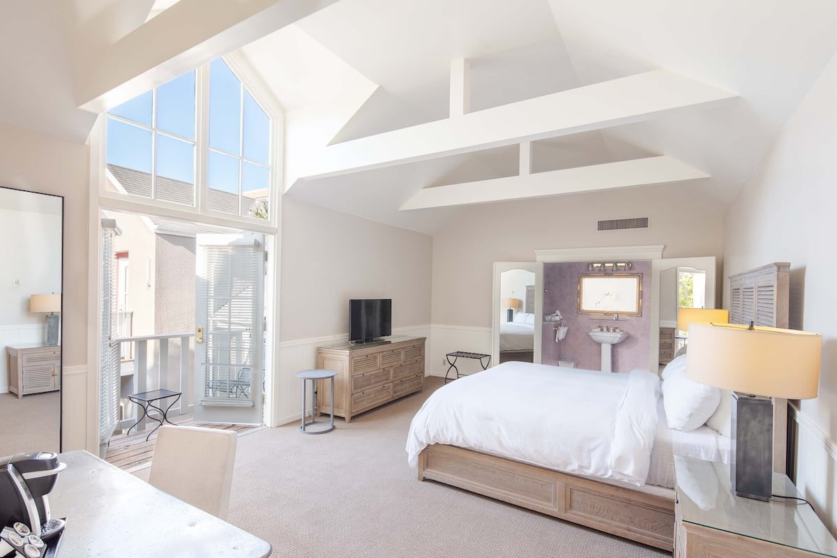 A hotel room with a large bed, a TV, a desk with a chair, a small table, and a balcony with a chair.