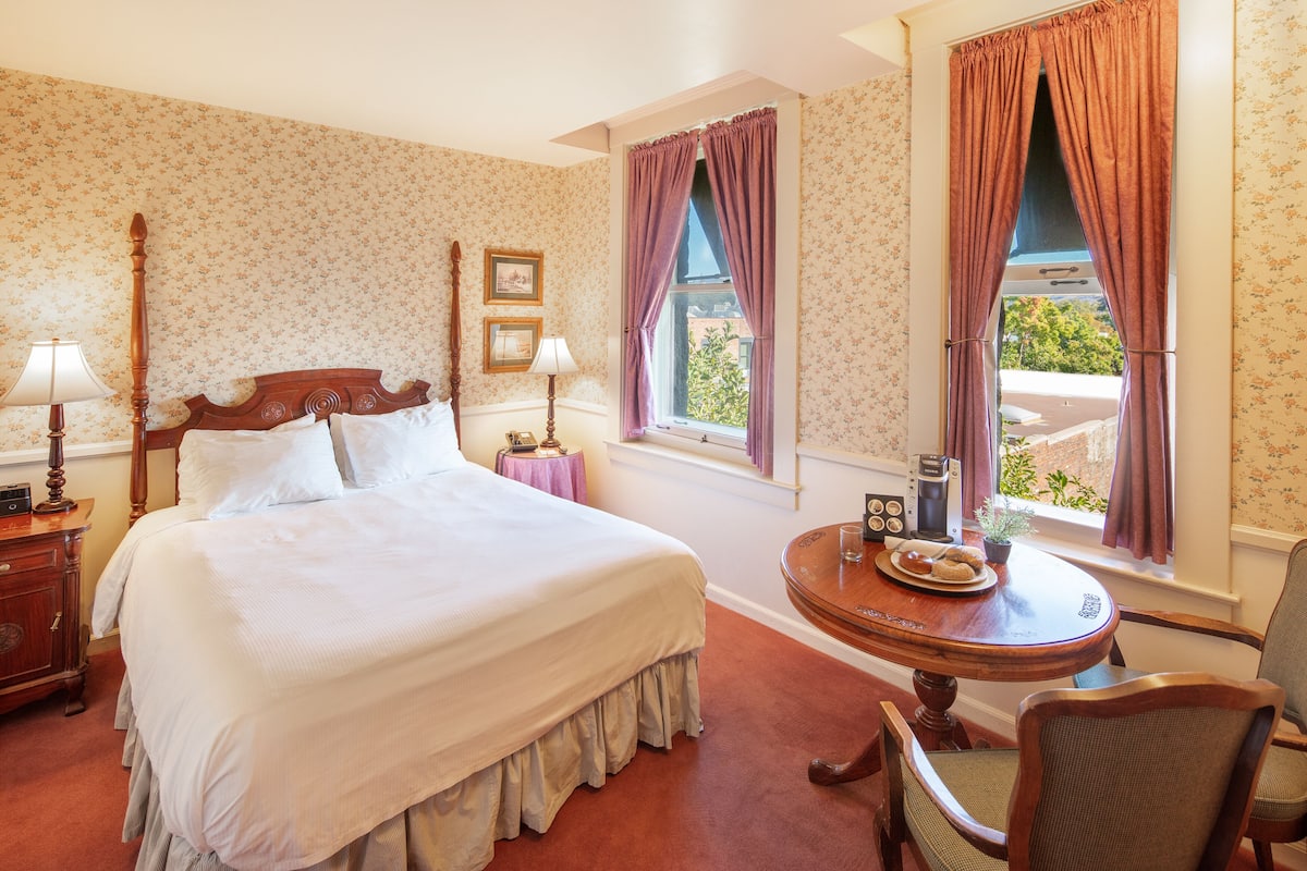 A neatly made bed with a wooden headboard, a bedside table with a lamp, a small round table with a plate of pastries, and a chair.
