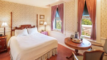 A neatly made bed with a wooden headboard, a bedside table with a lamp, a small round table with a plate of pastries, and a chair.