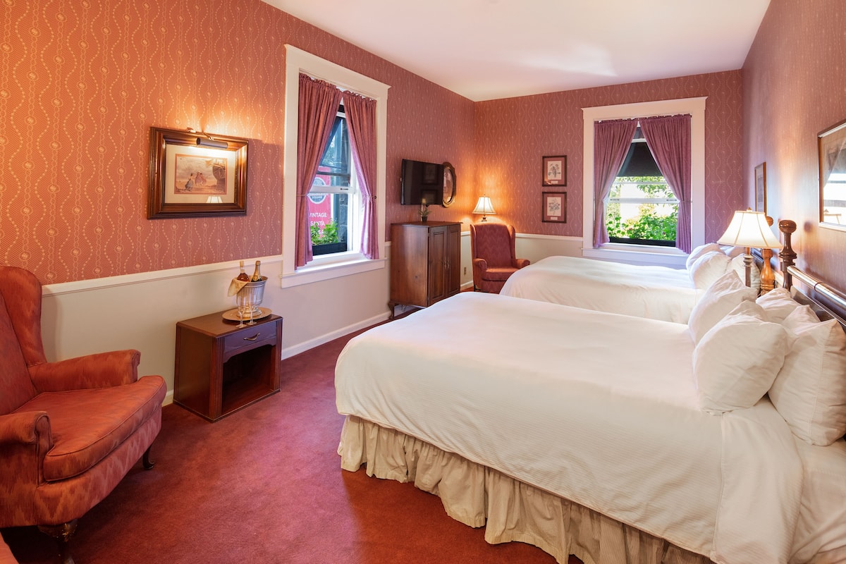 A hotel room with two beds, a red armchair, a small table with a lamp, and a television on a stand.