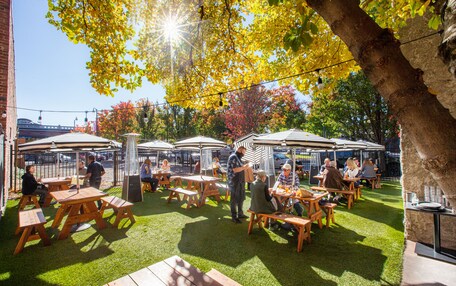 An outdoor patio area with wooden tables and benches, surrounded by trees and a brick wall.