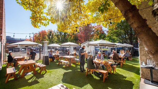 An outdoor patio area with wooden tables and benches, surrounded by trees and a brick wall. - Hotel La Rose (Santa Rosa)