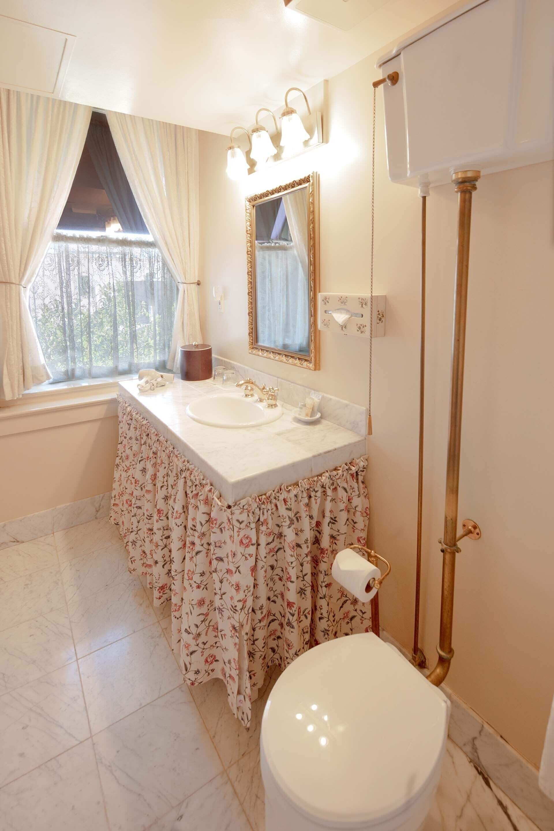 A bathroom with a marble floor, a white sink with a floral skirt, a toilet, and a window with curtains.