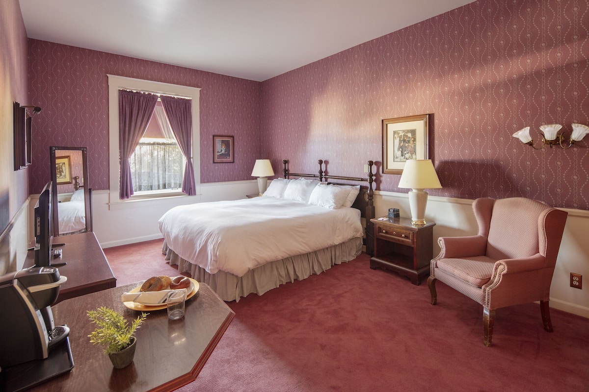 A hotel room with a bed, a desk with a computer, a chair, and a window with curtains.