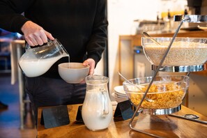 A person pouring milk into a bowl, with a jug and a bowl of cereal on a table.