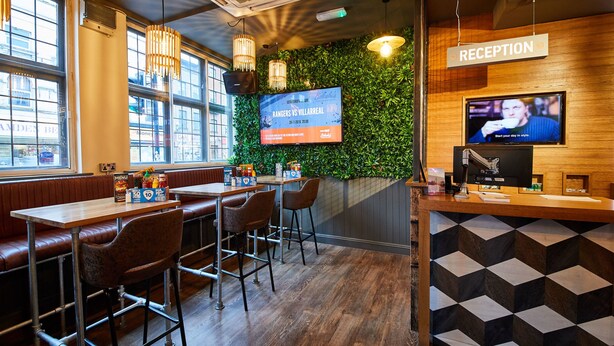 A restaurant with a reception area, wooden tables, bar stools, and a large TV screen displaying a menu. - St Christopher's Inn, Camden - Hostel (London)