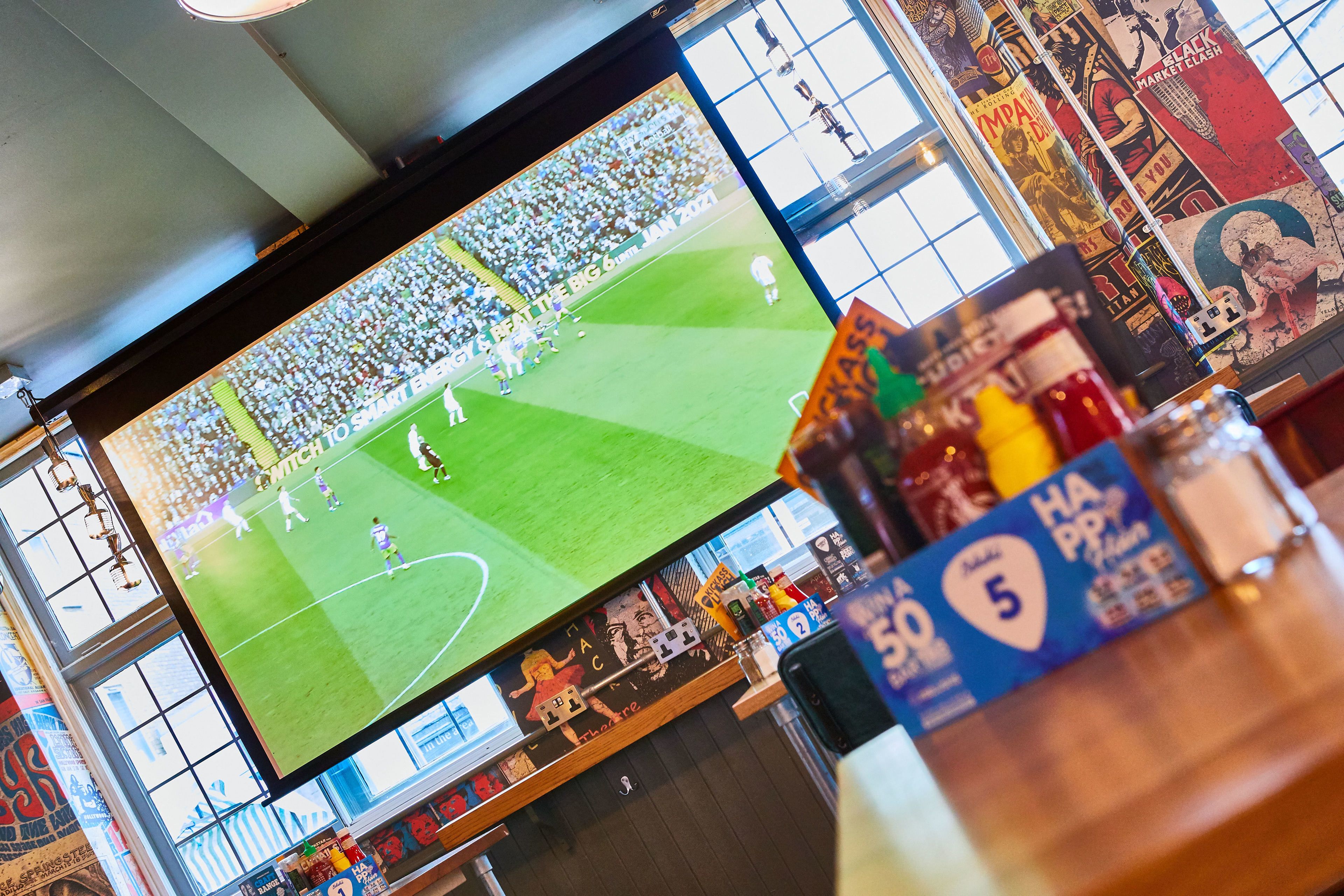 A large screen displaying a sports event in a venue with a high ceiling and large windows.