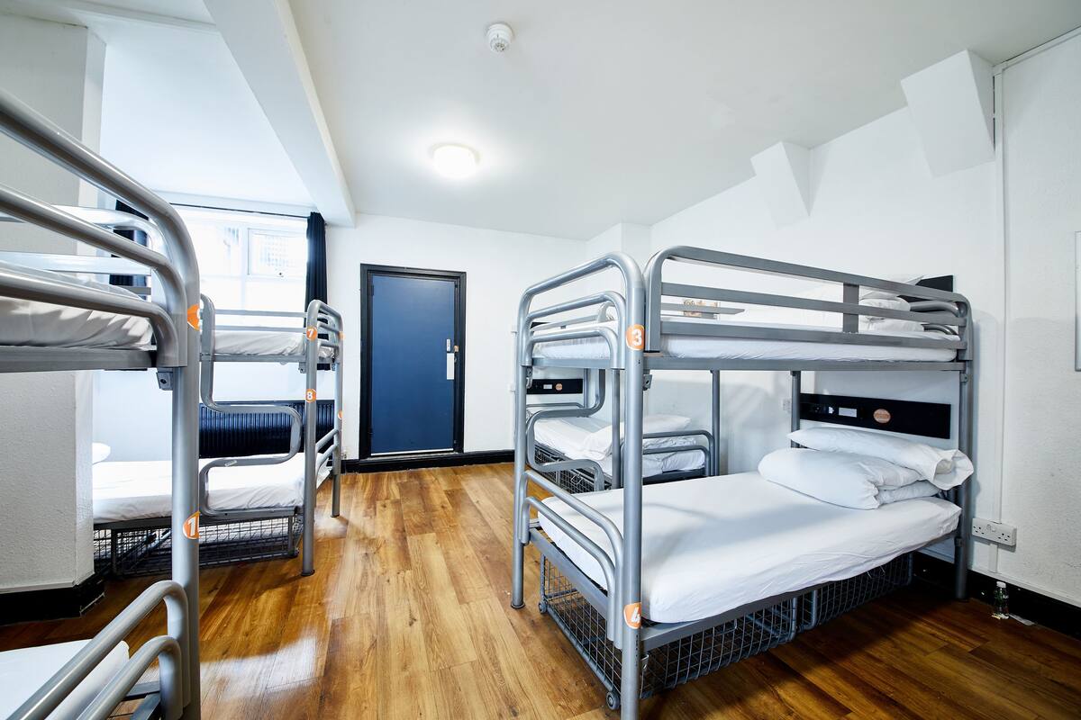 A dormitory room with bunk beds, a wooden floor, a door, and a window.