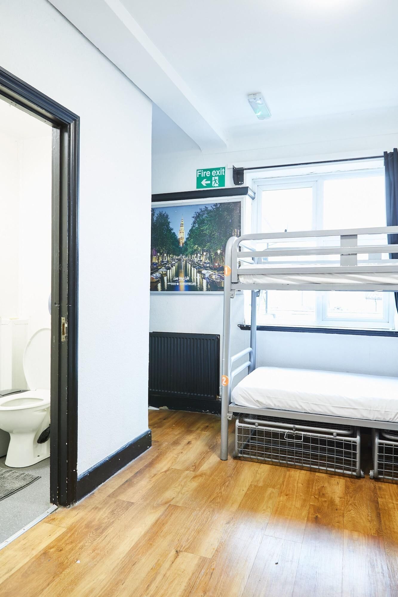 Un dormitorio tipo dormitorio con literas, un inodoro, un cartel de salida de incendios y un póster de un paisaje urbano.