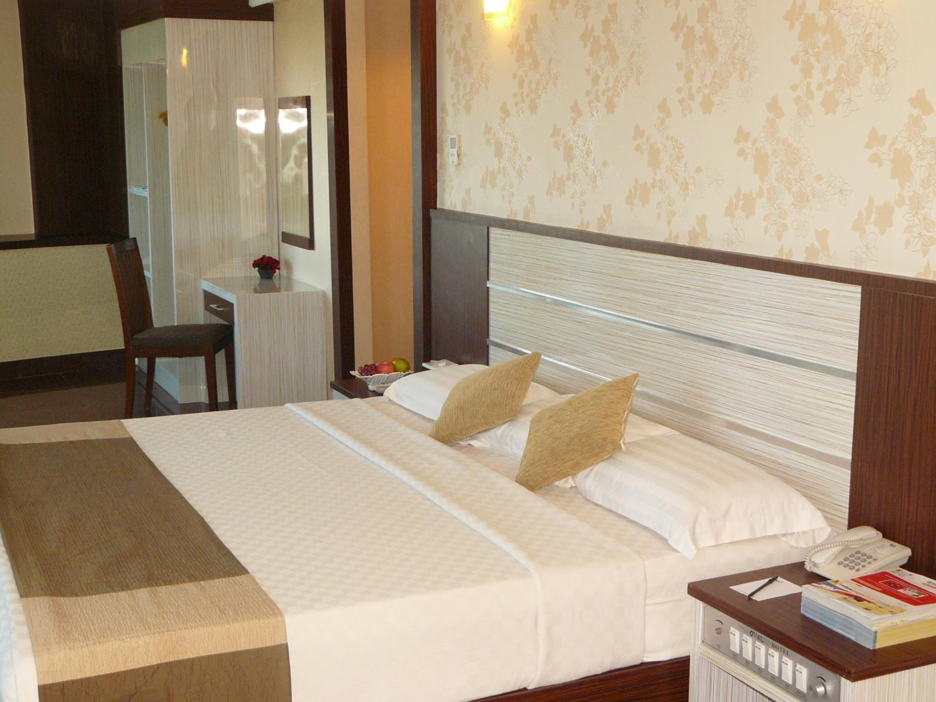 A hotel room with a large bed, a bedside table with a phone and books, a desk with a chair, and a mirror.