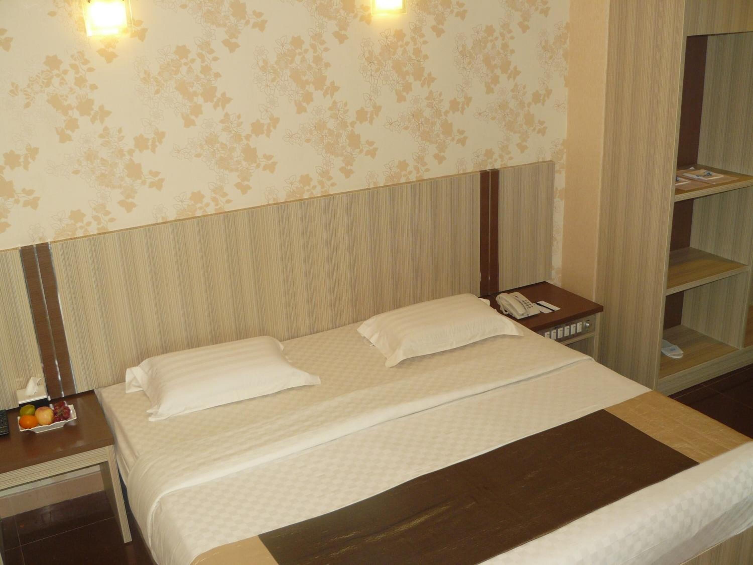 Una habitación de hotel con cama, mesita de noche, teléfono y una estantería con libros.