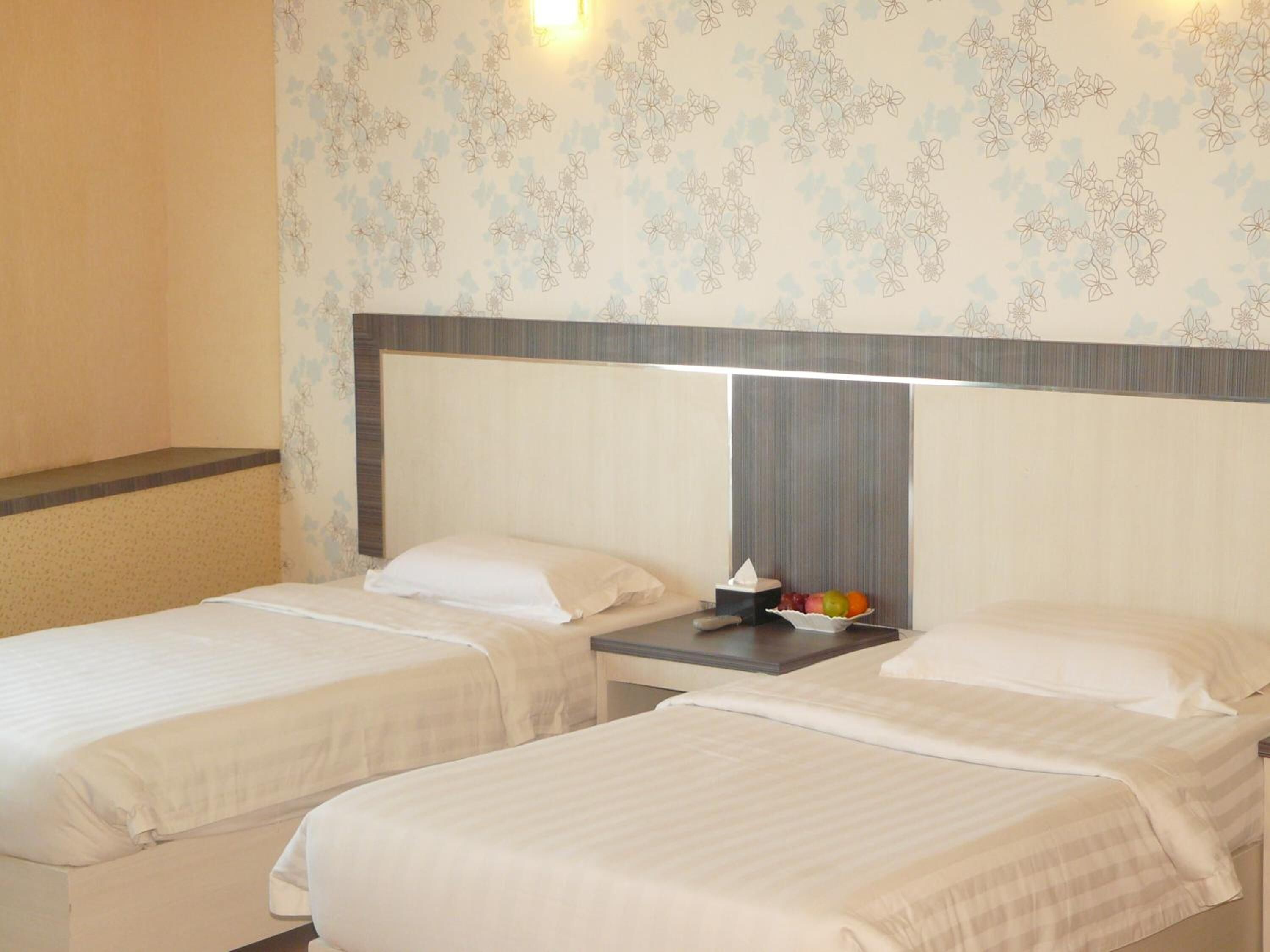 A hotel room with two beds, a nightstand with a tissue box and fruit, and floral wallpaper.
