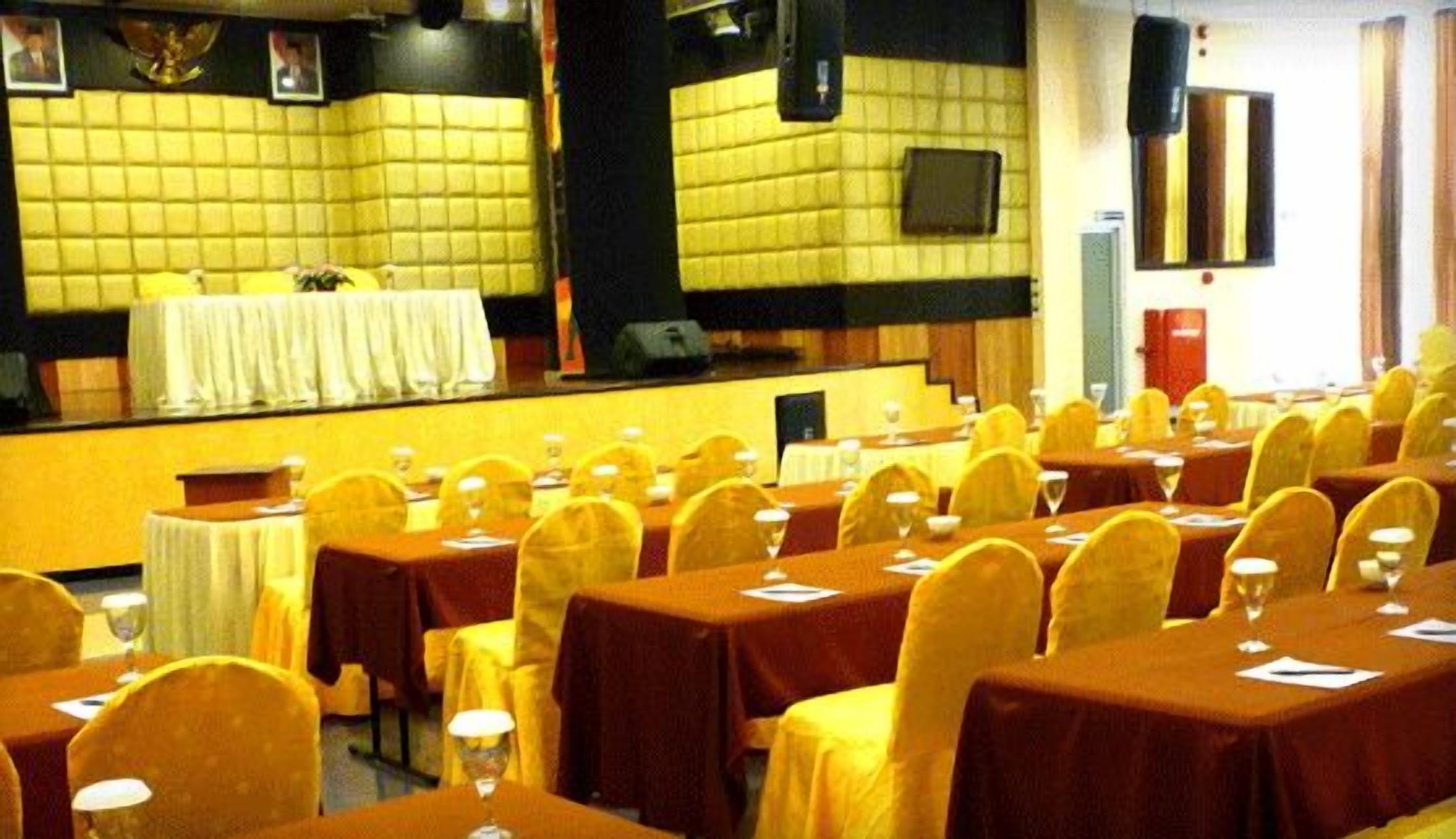 Un salón de conferencias con varias mesas y sillas, un escenario con un atril, y una pared amarilla con un diseño de cuadrícula.