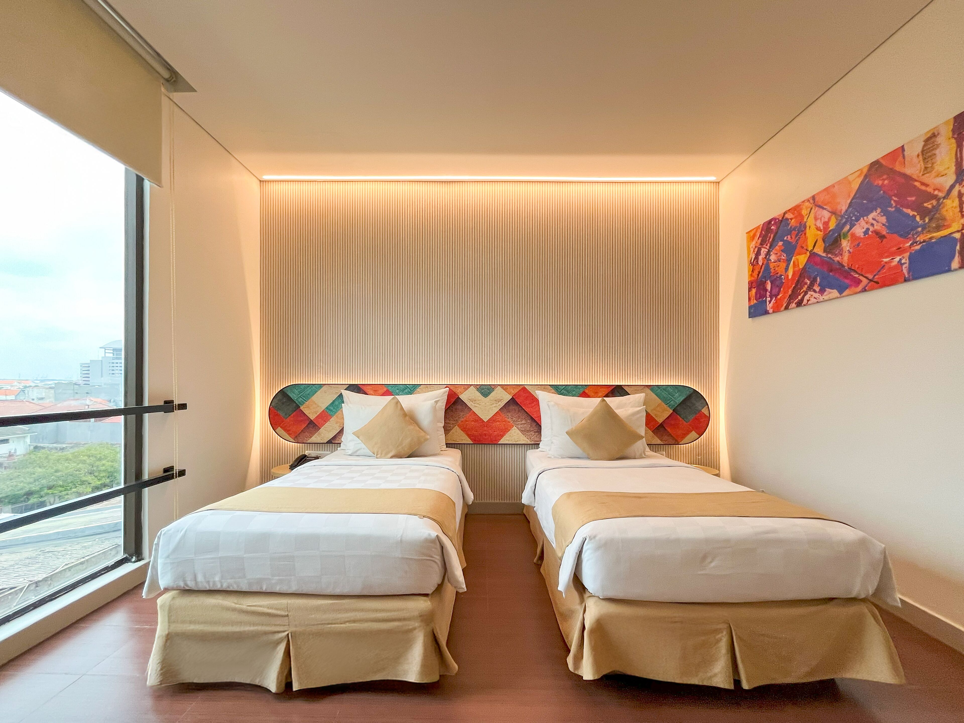 Habitació d'hotel amb dos llits, vista a la ciutat, un quadre abstracte i colorit, i un panell de paret a ratlles.