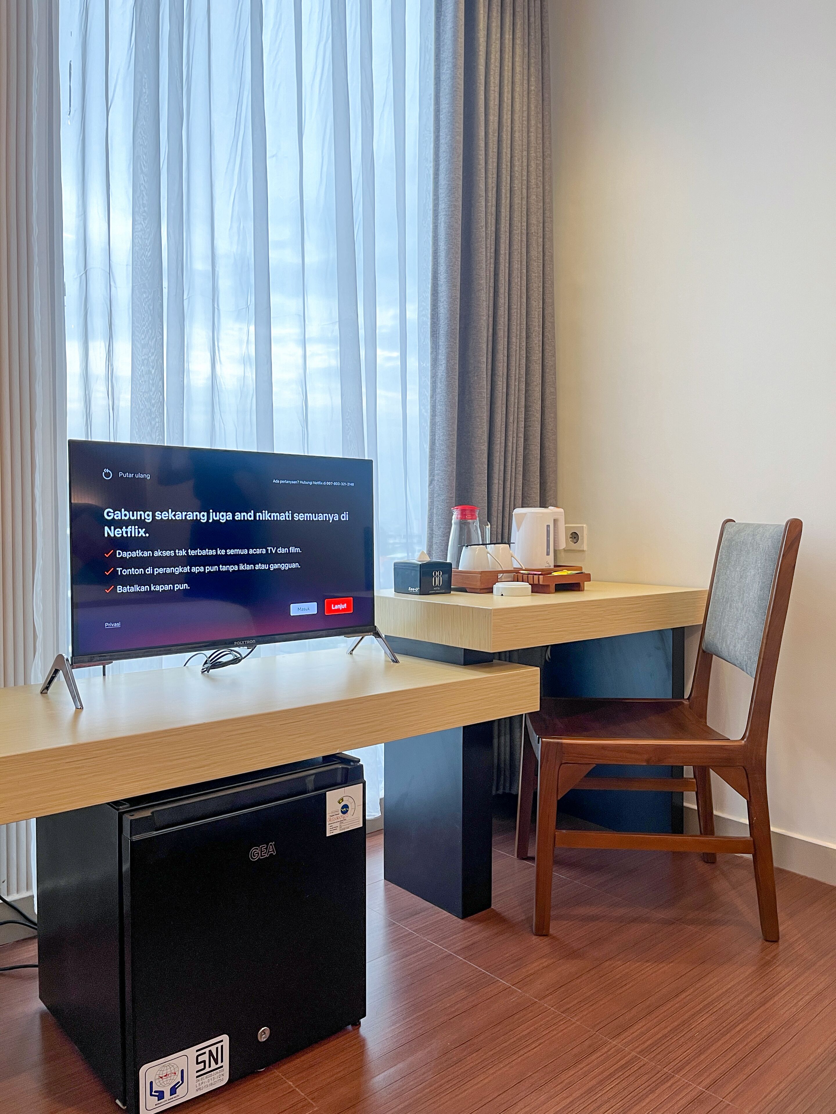 A hotel room with a flat-screen TV on a wooden desk, a small black refrigerator, a chair, and a window with curtains.