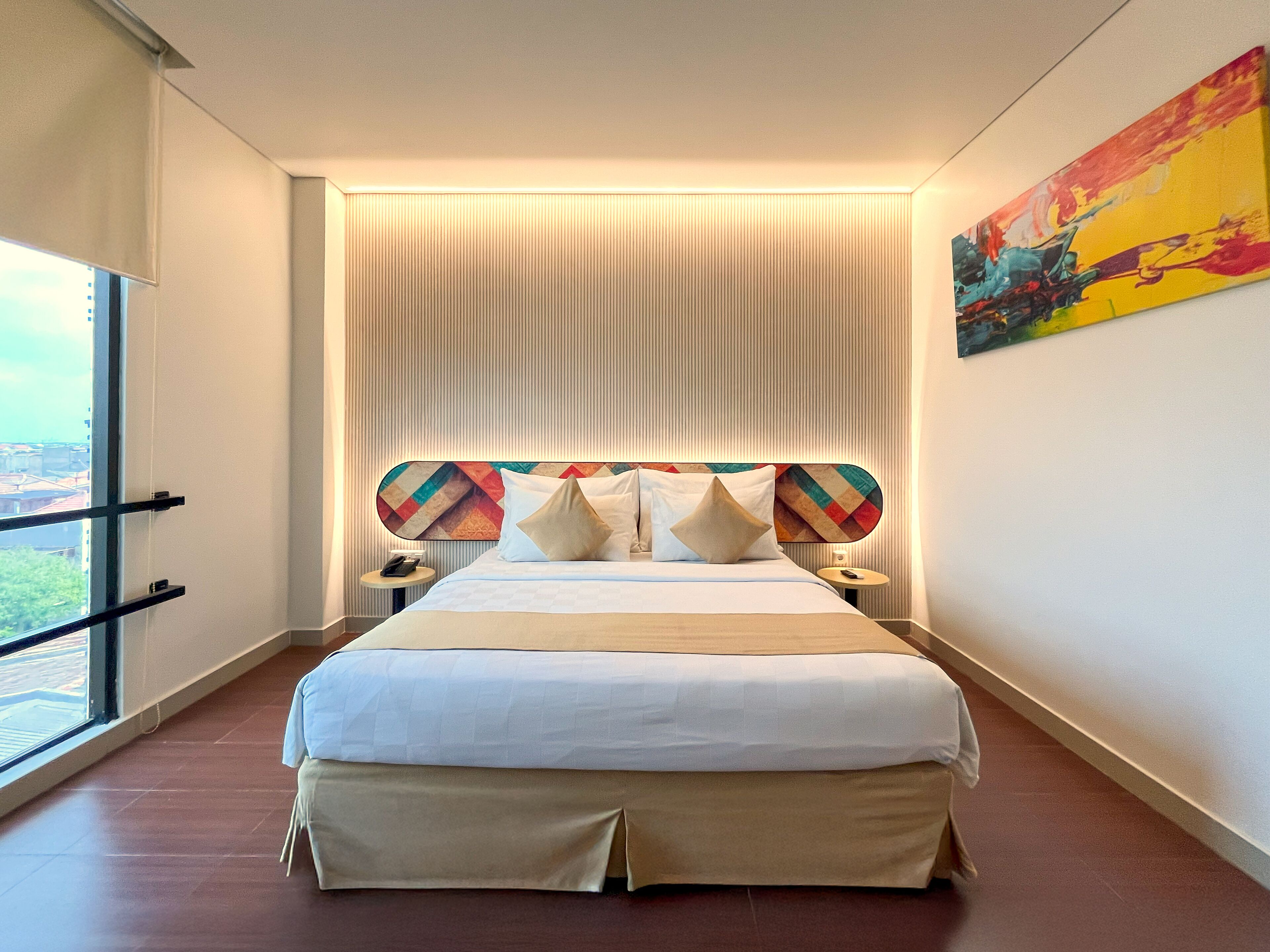 A modern hotel room with a large bed, a view through a window, and a colorful painting on the wall.