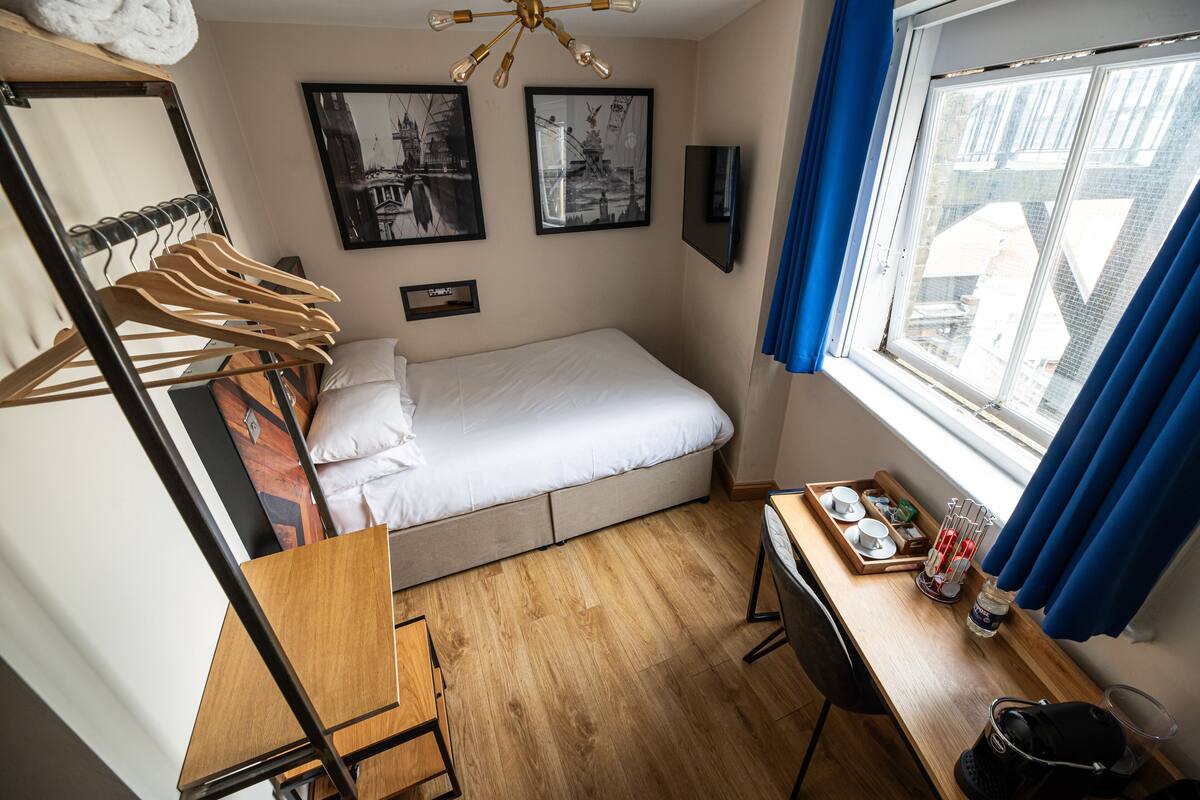 Quarto pequeno e bem iluminado, com uma cama, uma escrivaninha com uma bandeja de objetos, um cabideiro com roupas e uma janela com cortinas.