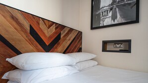 A neatly made bed with white linens and pillows, a wooden headboard with a chevron pattern, a black and white framed picture on the wall, and a small framed electrical outlet. - St Christopher's Inn Hammersmith (London)