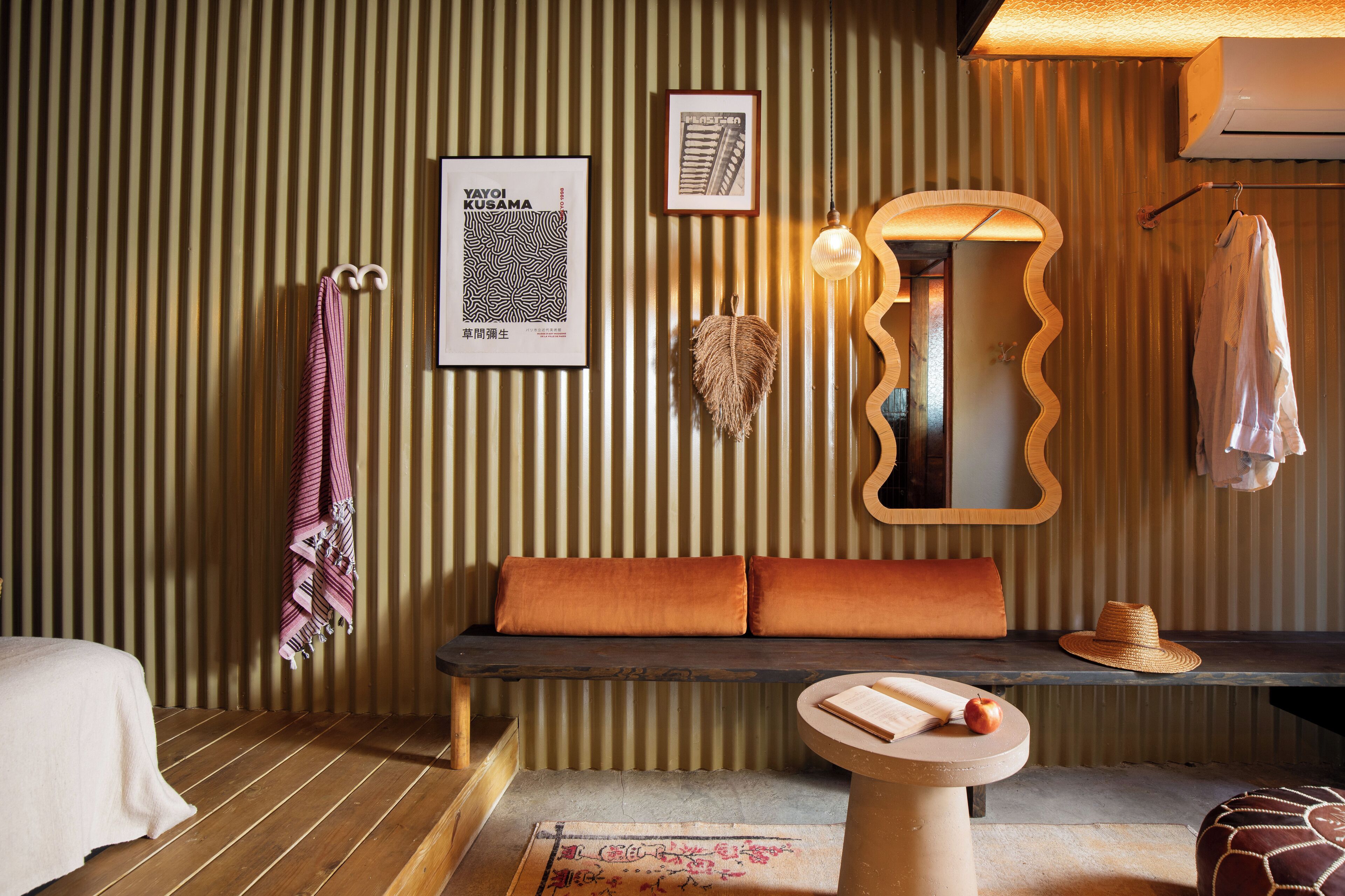 A room with a striped wall, a bench with cushions, a round table with an apple and a book, a hanging hat, and a towel on a hook.