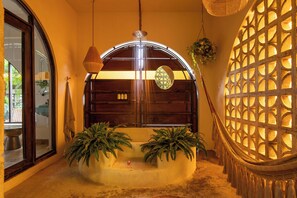 Une pièce comprenant un banc en bois aux lignes courbes, deux plantes en pot, un luminaire suspendu et un mur décoratif orné d’un motif en treillis.
