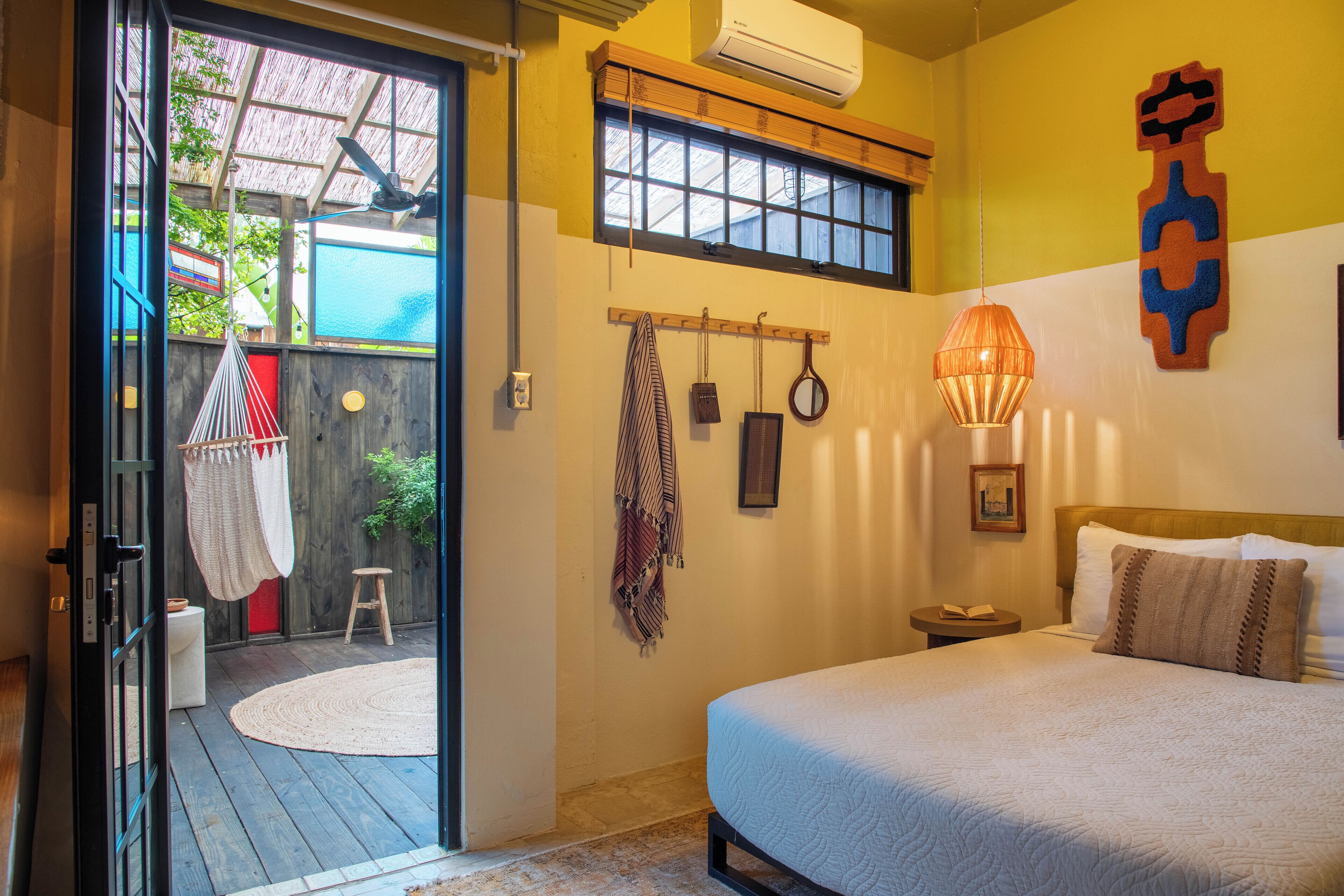 A bedroom with a bed, a bedside table, a hanging light fixture, a window with a view, and a door leading to an outdoor area.