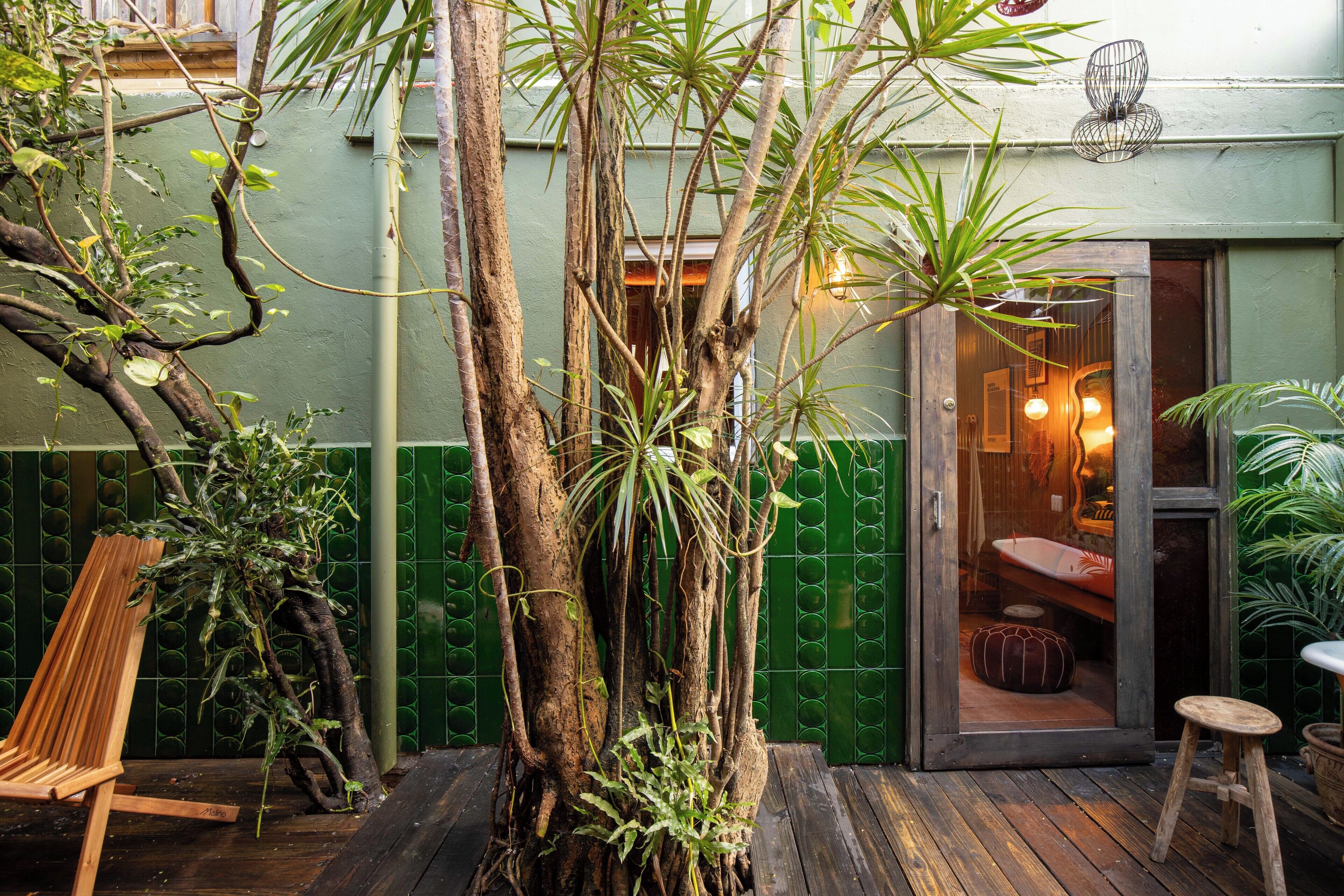 Een houten terras met een bankje en een klein krukje, omringd door weelderig groen en een grote boom.