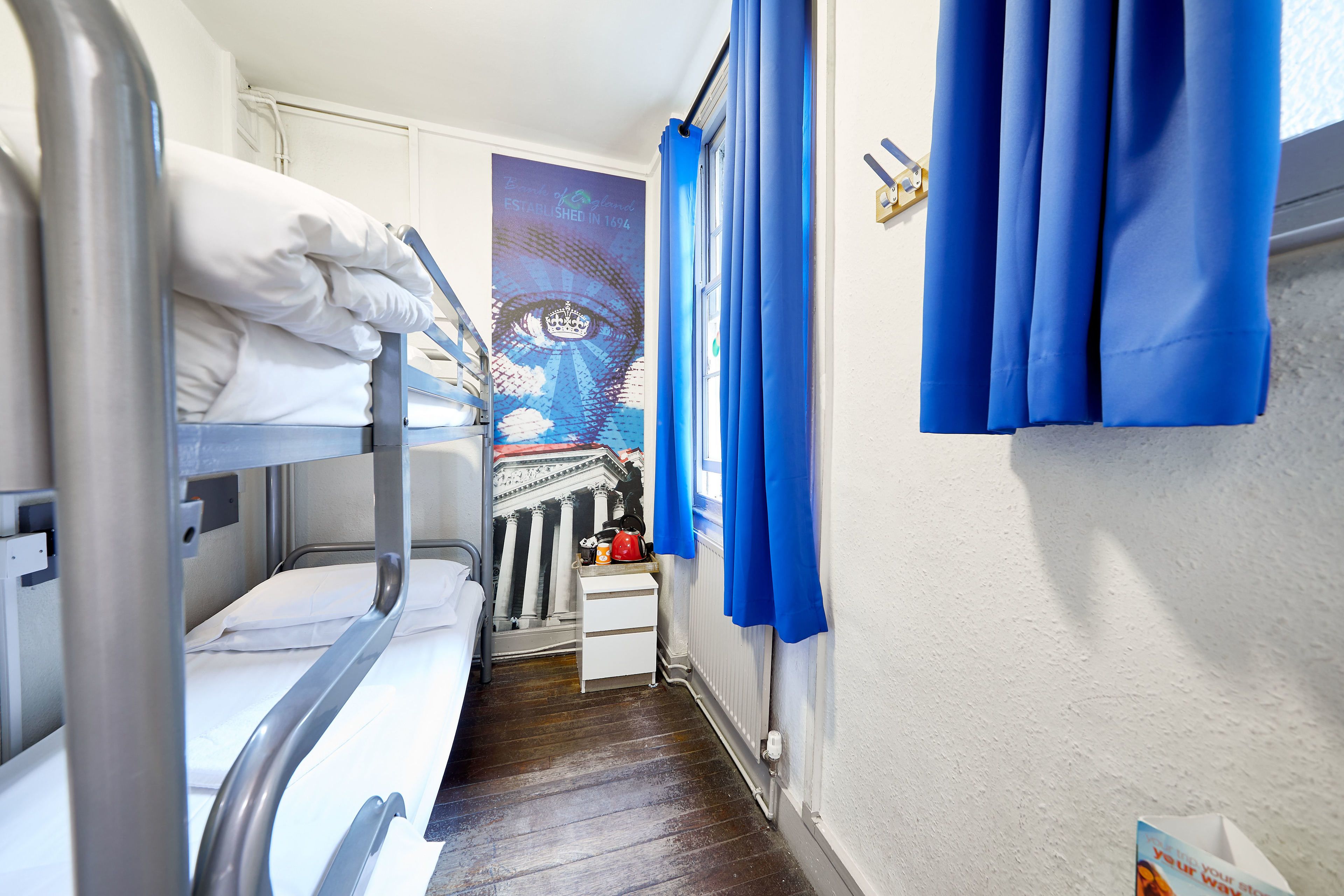A narrow corridor with bunk beds on one side and a small desk with a chair on the other. A large poster with an eye design is visible on the wall.