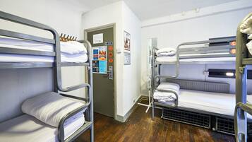 Habitación con literas, suelo de madera, barandillas metálicas y una puerta con un tablón de anuncios.