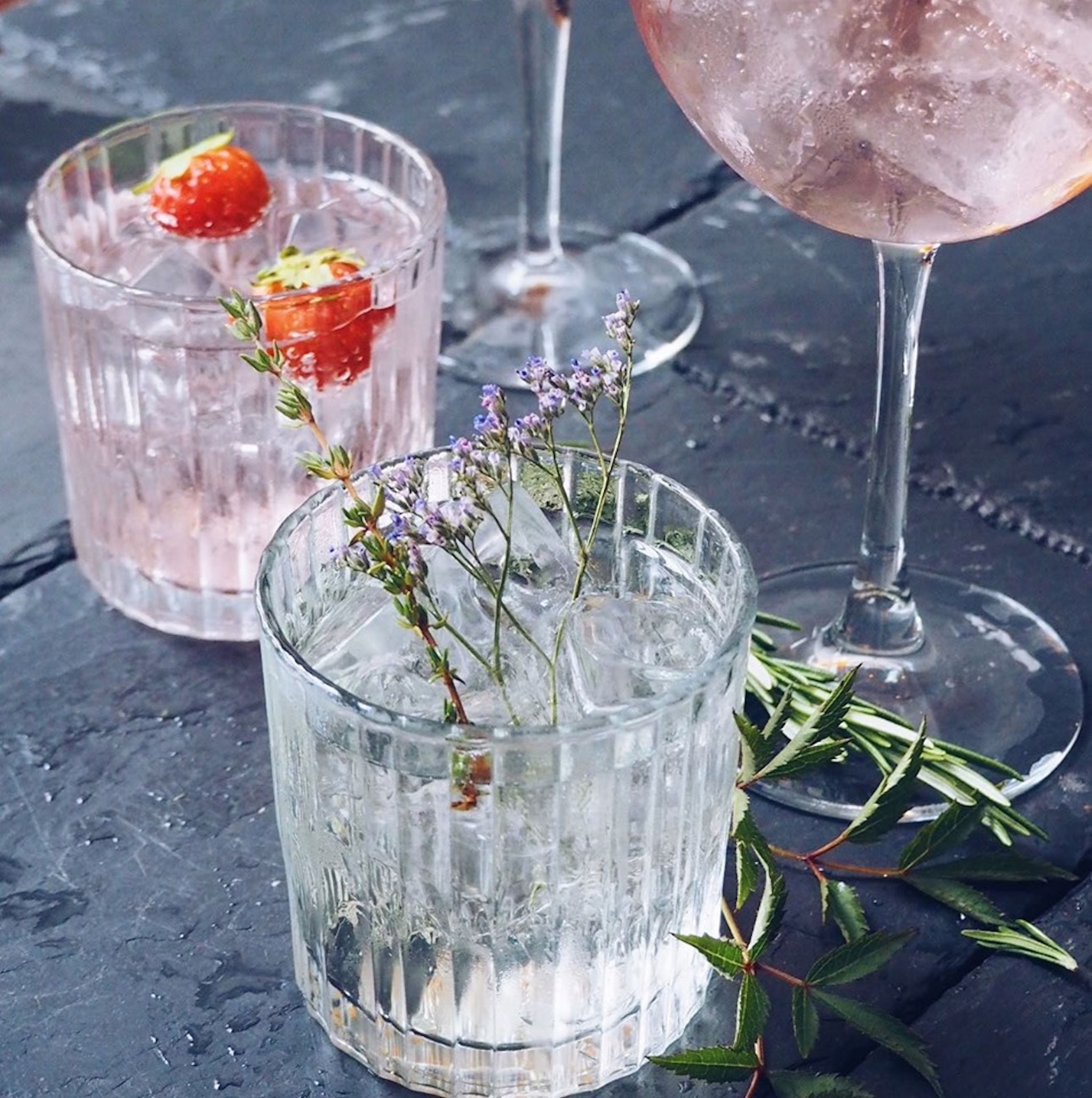 Two glasses of iced drinks with garnishes, one with a strawberry and mint, and the other with a purple flower.