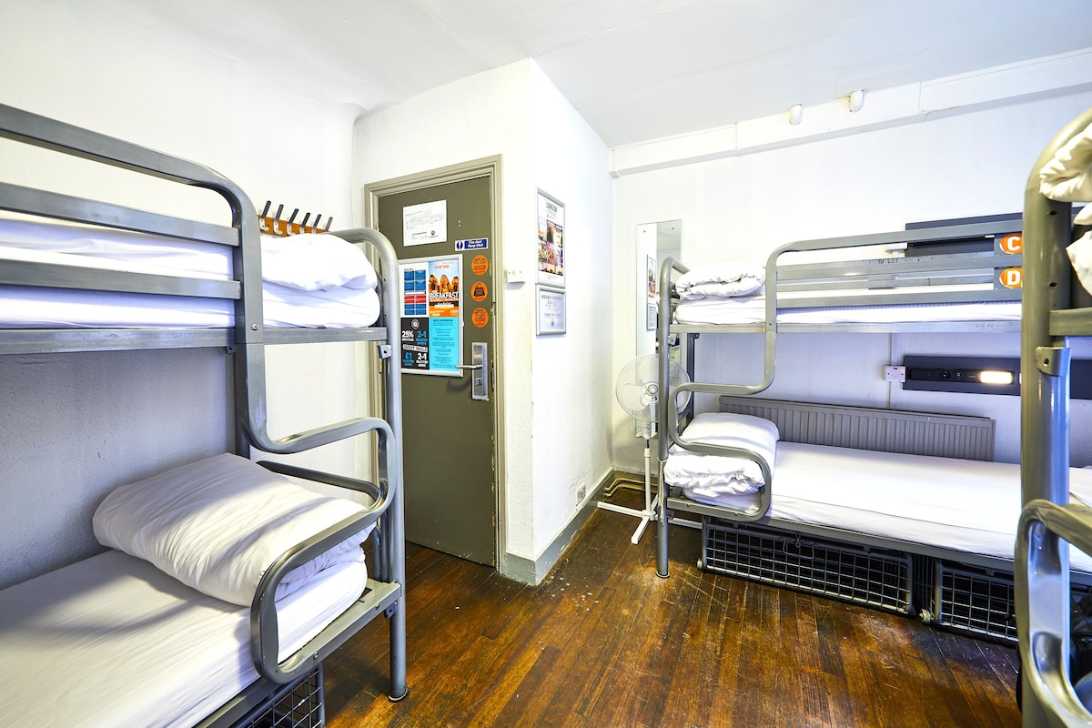 A bunk bed room with a wooden floor, a door with a notice, and a wall-mounted fan.