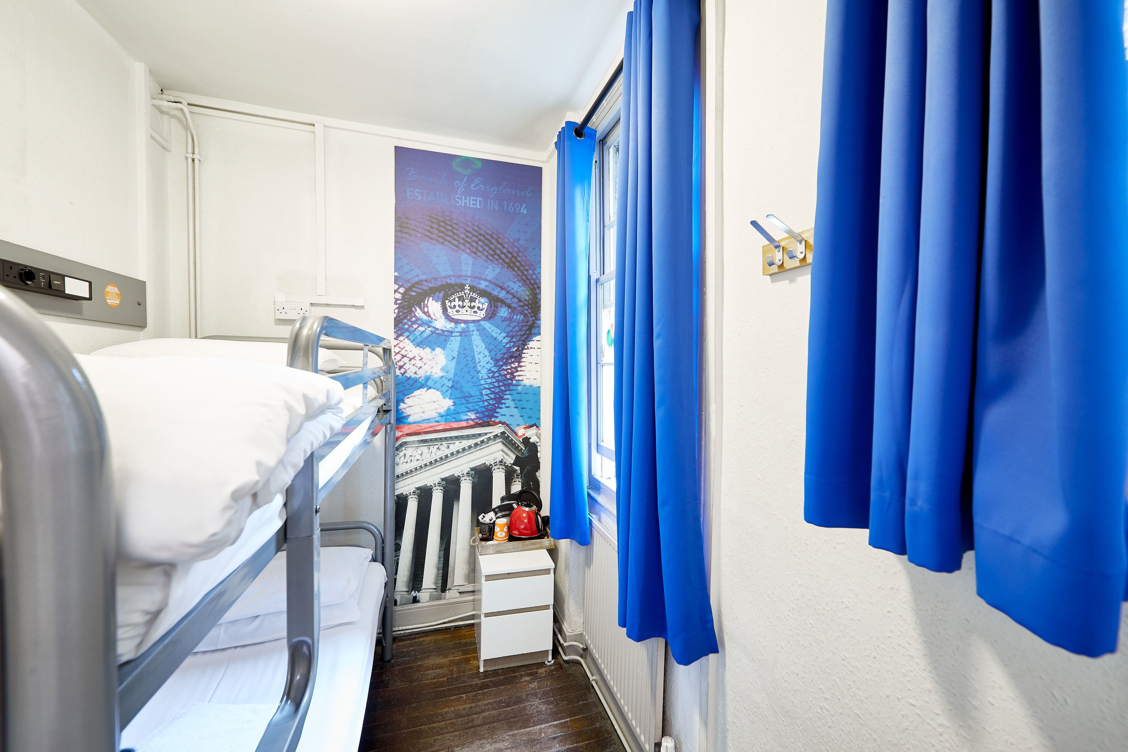 A narrow hotel room with bunk beds, a small bedside table with a kettle, a radiator, and a window with blue curtains.