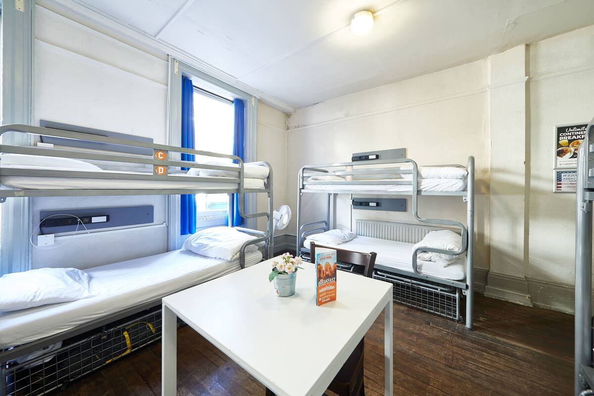 A dormitory room with bunk beds, a small table with a breakfast menu, and a window with blue curtains.