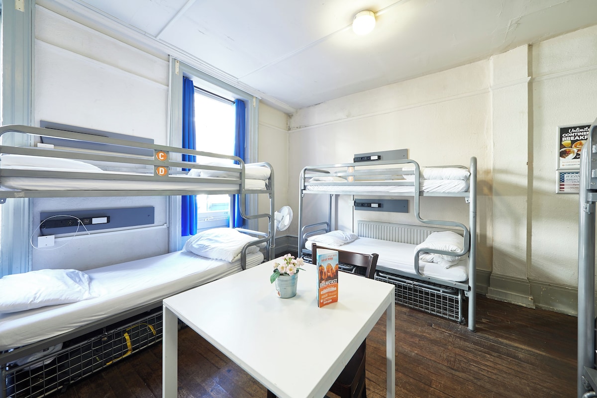 A dormitory room with bunk beds, a table with a sign, and a window with curtains.