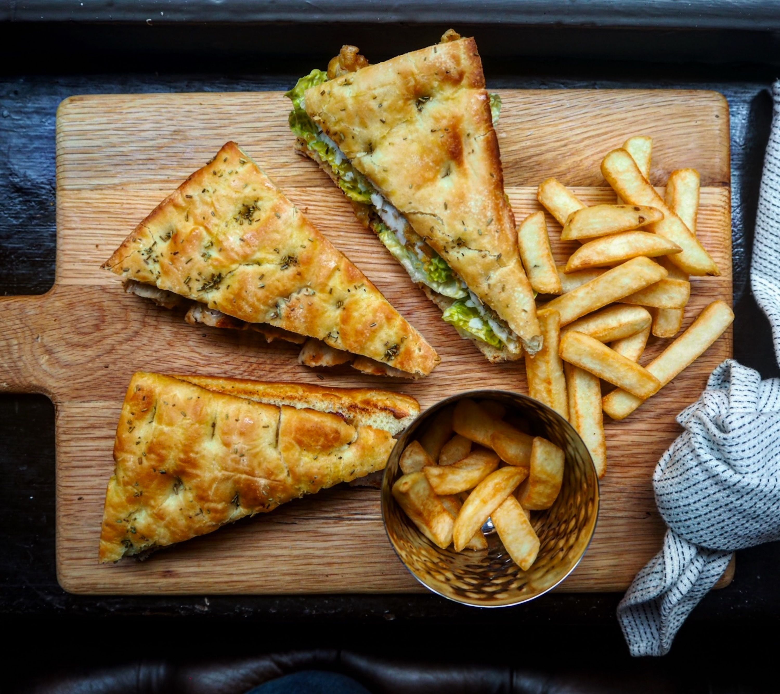 A wooden cutting board with a grilled sandwich, French fries, and a basket of fries.