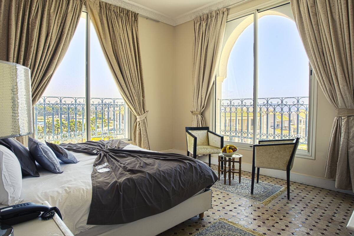 Ein Hotelzimmer mit einem großen Bett, einem Sessel, einem kleinen Tisch mit Obst und Blick auf einen Balkon mit Geländer.