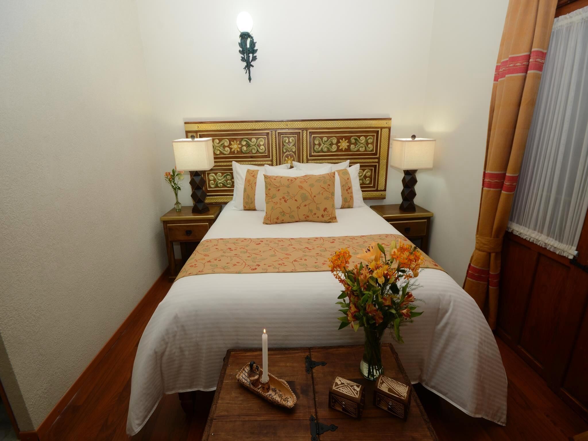 Habitación de hotel con una cama grande, dos mesitas de noche, una lámpara de pared, suelo de madera y una ventana con cortinas.